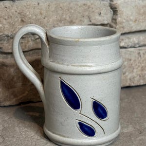 May include: A cylindrical ceramic mug with a speckled light gray finish and a single curved handle. The exterior features a debossed leaf pattern filled with a deep blue glaze, consisting of three distinct leaf shapes arranged on the side.