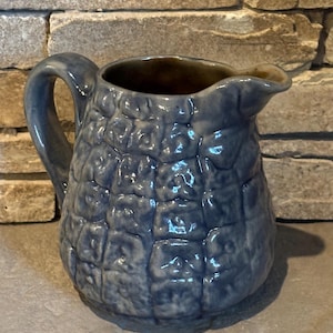 May include: A blue ceramic pitcher with a textured, cobblestone-like pattern. The pitcher has a handle and a spout for pouring. The interior is a darker brown color. The pitcher is likely used for serving beverages.