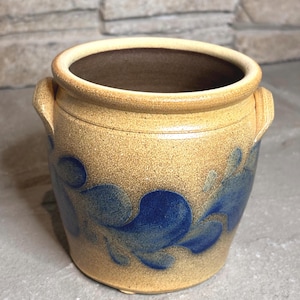 May include: A tan stoneware crock with a speckled texture and two small side handles. The exterior features a hand-painted cobalt blue floral vine pattern encircling the middle. The interior is a smooth, dark brown finish with a wide, open rim.