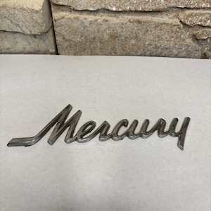 May include: A silver-colored Mercury emblem, featuring the brand name in a cursive font. The emblem is made of metal and is set against a white background. The emblem is likely for a car.