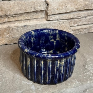 May include: A round ceramic ramekin featuring a deep navy blue glaze with a mottled cream-colored sponge-painted effect. The exterior of the dish has vertical fluted ridges around the circumference, creating a textured surface and a wide opening.
