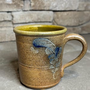 May include: A ceramic mug with a speckled brown glaze and a smooth, light green rim. The exterior features a hand-painted blue floral design on the side. The mug has a sturdy, curved handle and a cylindrical body with a horizontal embossed line.