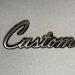 May include: A vintage, silver-toned "Custom" emblem with a cursive font. The emblem has a slightly distressed appearance, suggesting age or wear. The word is in a stylized script.