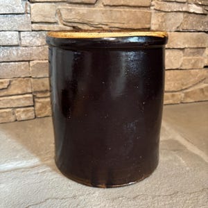 May include: A dark brown stoneware crock with a cylindrical shape. The antique jar has a slightly lighter brown rim and a smooth, glossy finish. The crock is likely used for food storage and has a rustic, vintage aesthetic.