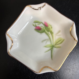 May include: A small, white, diamond-shaped dish with a scalloped gold rim. The dish features a hand-painted floral design with pink buds and green leaves. A decorative piece for trinkets or small items.