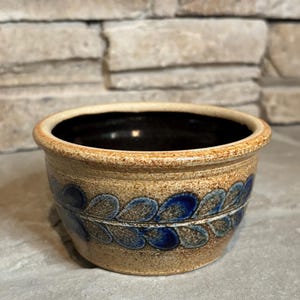 May include: A small, round ceramic bowl with a speckled tan and beige exterior featuring a horizontal band of blue, leaf-shaped patterns. The interior of the bowl is glazed in a dark, solid brown color, and the rim has a smooth, rounded edge.