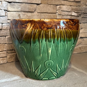May include: A ceramic planter with a wide, open top. The planter features a two-tone design, with a brown rim and a green body. The green section has a textured, carved pattern of vertical lines and stylized leaves.