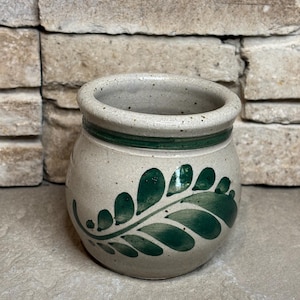 May include: A small, round, speckled ceramic pot with a light beige base color. The pot features a hand-painted dark green fern leaf design wrapping around the exterior and a solid dark green horizontal band encircling the upper rim.