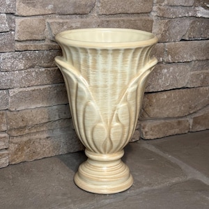 May include: A cream-colored ceramic vase with a fluted design and a pedestal base. The vase has two handles and a wide opening. The background is a stone wall.