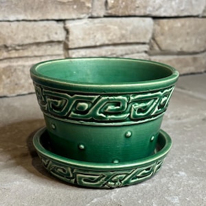 May include: A deep green ceramic plant pot with a matching saucer. The pot features a raised Greek key pattern around the upper rim and a series of small, evenly spaced raised dots around the lower body. The glaze has a subtle crackle finish.