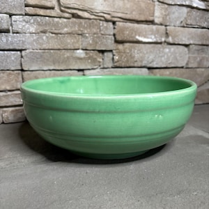 May include: A round, light green ceramic bowl with a smooth finish. The bowl has a simple design with two horizontal grooves near the top. The bowl is likely for serving food or as a decorative piece.