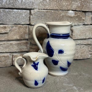 May include: A set of two off-white ceramic pitchers featuring a glossy cobalt blue floral pattern. The larger pitcher has a tall neck and a curved handle, while the smaller pitcher is rounded with a wide spout and a matching handle design.