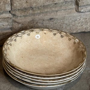 May include: A stack of four shallow ceramic bowls featuring a cream-colored base with a distinct crackle glaze finish. Each bowl is decorated with a dark brown geometric and leaf-patterned border along the rim, creating a cohesive set of dinnerware.
