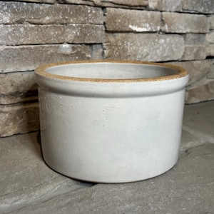 May include: A round, off-white ceramic crock with a light brown rim. The crock is cylindrical with a smooth, matte finish. It sits on a gray stone surface against a textured stone wall.