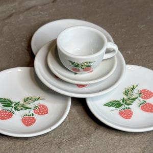 May include: A white ceramic tea set featuring a cup, saucer, and plates, all adorned with a strawberry motif. The strawberries are red with green leaves. The set includes a cup, a small saucer, and three plates.