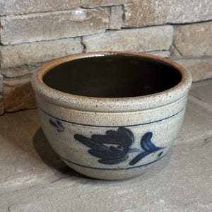 May include: A round ceramic bowl with a speckled beige exterior and a dark brown interior. The exterior features a hand-painted dark blue floral motif centered between two thin horizontal blue lines encircling the body of the bowl.