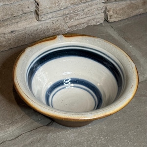 May include: A handmade ceramic bowl with a white interior and two dark blue bands. The rim and exterior are a warm brown. The bowl has a slightly irregular shape, suggesting it is hand-crafted. The bowl is likely for serving or display.