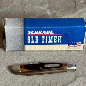 May include: A Schrade Old Timer pocket knife with a brown handle and silver accents, next to its blue and white box. The box features the brand name and the American flag, indicating it's made in the USA.