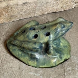 May include: A ceramic frog-shaped flower frog with a mottled green and blue glaze. The sculpture features a series of circular holes across the back, designed to hold flower stems in place, with a smooth, rounded base and a textured, glossy finish.