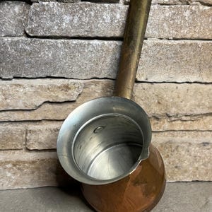 May include: A copper and brass Turkish coffee pot with a long handle and a silver-colored interior. The pot has a rounded base and a spout for easy pouring. The handle has a hole for hanging.
