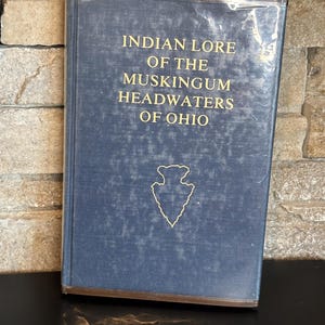 May include: A hardcover book titled "Indian Lore of the Muskingum Headwaters of Ohio" with gold lettering and a gold arrowhead graphic on a blue cover. The book is wrapped in clear plastic.