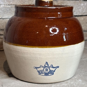 May include: A two-toned stoneware crock featuring a dark brown glazed upper section and a cream-colored lower body. A blue stamped graphic of a crown with the number 2 inside is centered on the front. The crock has a short, thick neck and a lid.