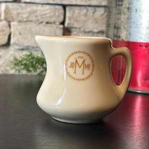 May include: Cream-colored ceramic creamer with a curved body and a handle. The creamer has a gold rim and a gold emblem that reads "AND JD MHE" in a circular design. The creamer is on a black surface.