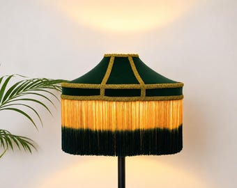 May include: A dark green velvet lampshade featuring gold braided trim along the panels and a long, two-toned fringe that transitions from a warm golden yellow at the top to a deep dark green at the bottom edge, illuminated from within.