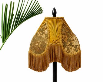 May include: A vintage-style lampshade with a mustard yellow and gold floral lace design. The lampshade has a fringed bottom and is mounted on a black pole. A green palm frond is in the background.