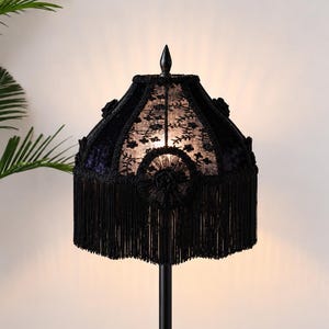 May include: A black lamp shade featuring intricate floral lace panels, decorative fabric rosettes, and a long, dense fringe trim along the bottom edge. The shade is topped with a pointed finial and emits a soft, warm glow from within.
