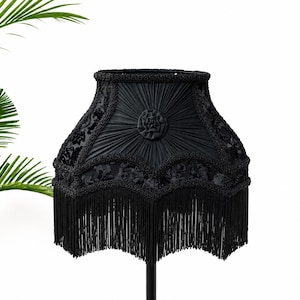 May include: A black lampshade with a decorative design and fringe. The lampshade features a textured pattern, a beaded trim, and a central rosette. The fringe hangs from the bottom edge. The lampshade is on a black stand.