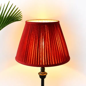 Puede incluir: Una lámpara de mesa con una pantalla de tela plisada de color rojo vibrante y forma cónica. La pantalla está montada sobre una base de metal oscura y delgada. La lámpara emite un brillo cálido y suave desde el interior, iluminando la tela roja.