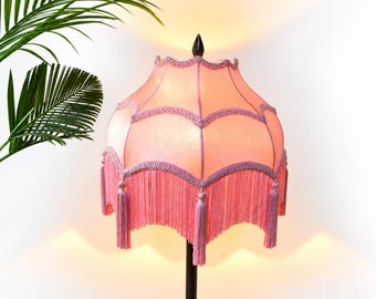 Op de afbeelding: Een klokvormige lampenkap in een zachte perzikkleur, voorzien van geschulpte randen afgezet met een gevlochten lavendelkleurig koord. De onderrand is versierd met lange, bijpassende lavendelkleurige franjes en een donkere knop op het middelpunt.
