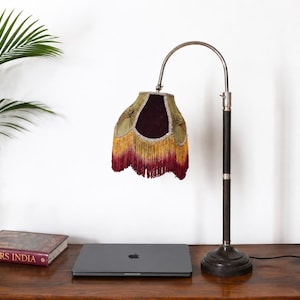 Peut inclure: Une lampe de table avec une base en métal sombre et un col incurvé. L'abat-jour est recouvert de tissu vert olive avec un panneau en velours violet foncé, orné de broderies dorées et d'une longue frange superposée dans des tons jaune et rouge.