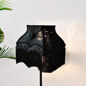 Pode incluir: Um abajur preto com silhueta quadrada, painéis de tecido plissado, um detalhe de roseta central, acabamento em renda intrincada ao longo da borda inferior e uma franja longa e densa que pende da base do abajur para criar textura.