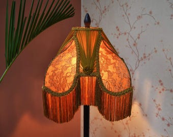 Op de afbeelding: Een klokvormige lampenkap met een warme oranje kanten stof met bloemmotief. De kap is afgewerkt met olijfgroene gevlochten biezen langs de naden en randen, en voorzien van een lange, dichte franjerand in een bijpassende olijfgroene kleur.