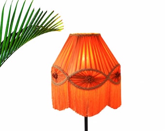 May include: A vibrant orange pleated lampshade featuring a decorative scalloped trim with circular braided accents and a long fringe border. The lampshade has a conical shape and is illuminated from within, casting a warm glow across the fabric.
