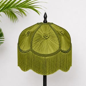 May include: A bell-shaped lampshade in olive green fabric featuring intricate radial pleating, decorative fabric rosettes at the top of each panel, and a long, matching green fringe trim along the scalloped bottom edge, topped with a black finial.