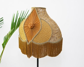 May include: A vintage lampshade in shades of gold and tan, featuring a decorative floral pattern and a fringed bottom. The lampshade has a unique, layered design with a central, textured accent. A green plant is in the background.