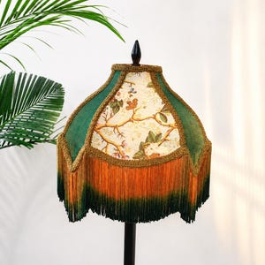 May include: A bell-shaped lamp shade featuring panels of cream fabric with a floral branch pattern alternating with dark green velvet panels. The shade is trimmed with gold braided cord and finished with a long, two-toned orange and green fringe.