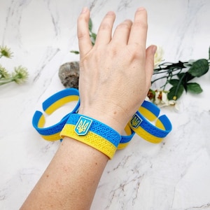 Può includere: Un set di braccialetti in silicone flessibile con i colori blu e giallo della bandiera ucraina. Ogni braccialetto presenta un motivo geometrico strutturato e un emblema centrale che mostra il simbolo giallo del tridente ucraino.