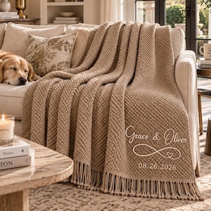 Op de afbeelding: Een beige, getextureerde gebreide deken met franjes, voorzien van de geborduurde witte tekst Grace & Oliver, een oneindigheidssymbool met een klein hartje en de datum 26-08-2026, gedrapeerd over een lichtgekleurde bank in een woonkamer.