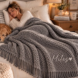 Può includere: Una coperta grigia in maglia grossa con un motivo testurizzato a pallini e lunghe frange sul bordo inferiore. La coperta presenta il testo ricamato in corsivo bianco "Miles" seguito da un piccolo simbolo a forma di cuore nell'angolo in basso a destra.