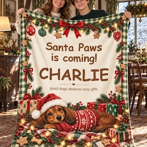 Könnte beinhalten: Eine cremefarbene Wohndecke mit grün-weißem Karomuster am Rand, zeigt einen braunen Dackel im roten Pullover mit Weihnachtsmütze, umgeben von Geschenken, Tannenzweigen und dem Text Santa Paws is coming! CHARLIE.