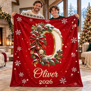 Könnte beinhalten: Eine rote Fleecedecke mit einem großen, goldenen Buchstaben O in der Mitte, umgeben von einem festlichen Kranz aus Tannenzweigen, roten Beeren, Tannenzapfen und einer roten Schleife. Der Name Oliver und das Jahr 2026 stehen in Weiß.