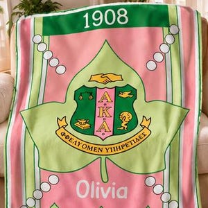 K&ouml;nnte beinhalten: Eine rosa-gr&uuml;ne Fleecedecke mit dem Alpha Kappa Alpha Wappen, der Jahreszahl 1908 und dem Namen Olivia. Das Design zeigt ein gro&szlig;es Efeublatt, wei&szlig;e kreisf&ouml;rmige Akzente und den Schriftzug Alpha Kappa Alpha am unteren Rand.