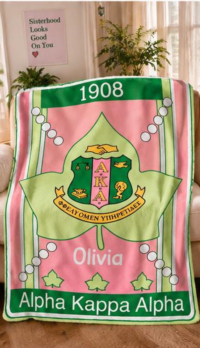 K&ouml;nnte beinhalten: Eine rosa-gr&uuml;ne Fleecedecke mit dem Alpha Kappa Alpha Wappen, der Jahreszahl 1908 und dem Namen Olivia. Das Design zeigt ein gro&szlig;es Efeublatt, wei&szlig;e kreisf&ouml;rmige Akzente und den Schriftzug Alpha Kappa Alpha am unteren Rand.