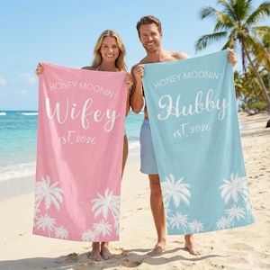 May include: Two beach towels, one pink and one light blue, with white text and palm tree designs. The pink towel reads "Honey Moonin' Wifey est. 2026". The blue towel reads "Honey Moonin' Hubby est. 2026".