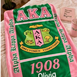 K&ouml;nnte beinhalten: Eine rechteckige Fleecedecke mit einem gr&uuml;nen Mittelteil, dem rosa Schriftzug AKA, einem zentralen Wappen und der Jahreszahl 1908. Der Name Olivia ist darunter in Wei&szlig; gedruckt. Die Decke hat rosa Seitenr&auml;nder mit dem Text Alpha Kappa Alpha.