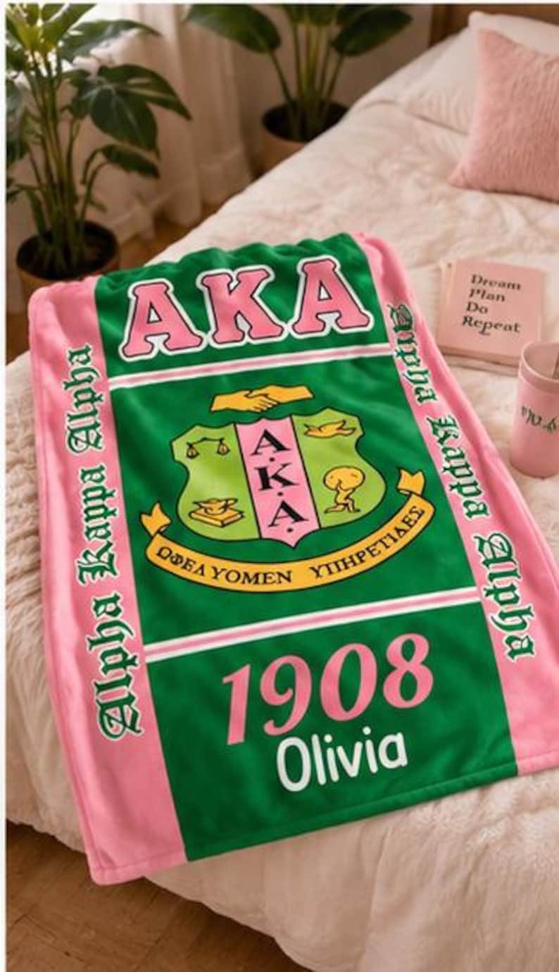 K&ouml;nnte beinhalten: Eine rechteckige Fleecedecke mit einem gr&uuml;nen Mittelteil, dem rosa Schriftzug AKA, einem zentralen Wappen und der Jahreszahl 1908. Der Name Olivia ist darunter in Wei&szlig; gedruckt. Die Decke hat rosa Seitenr&auml;nder mit dem Text Alpha Kappa Alpha.
